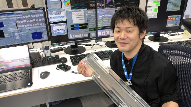 Ichiro Inoue working in the control room of SACLA (Japanese X-ray free-electron laser facility), where he is controlling the machine to generate twin XFEL pulses. He is holding an X-ray mirror for focusing the X-ray beam to micrometer size. (Credit: SACLA / IFJ PAN)